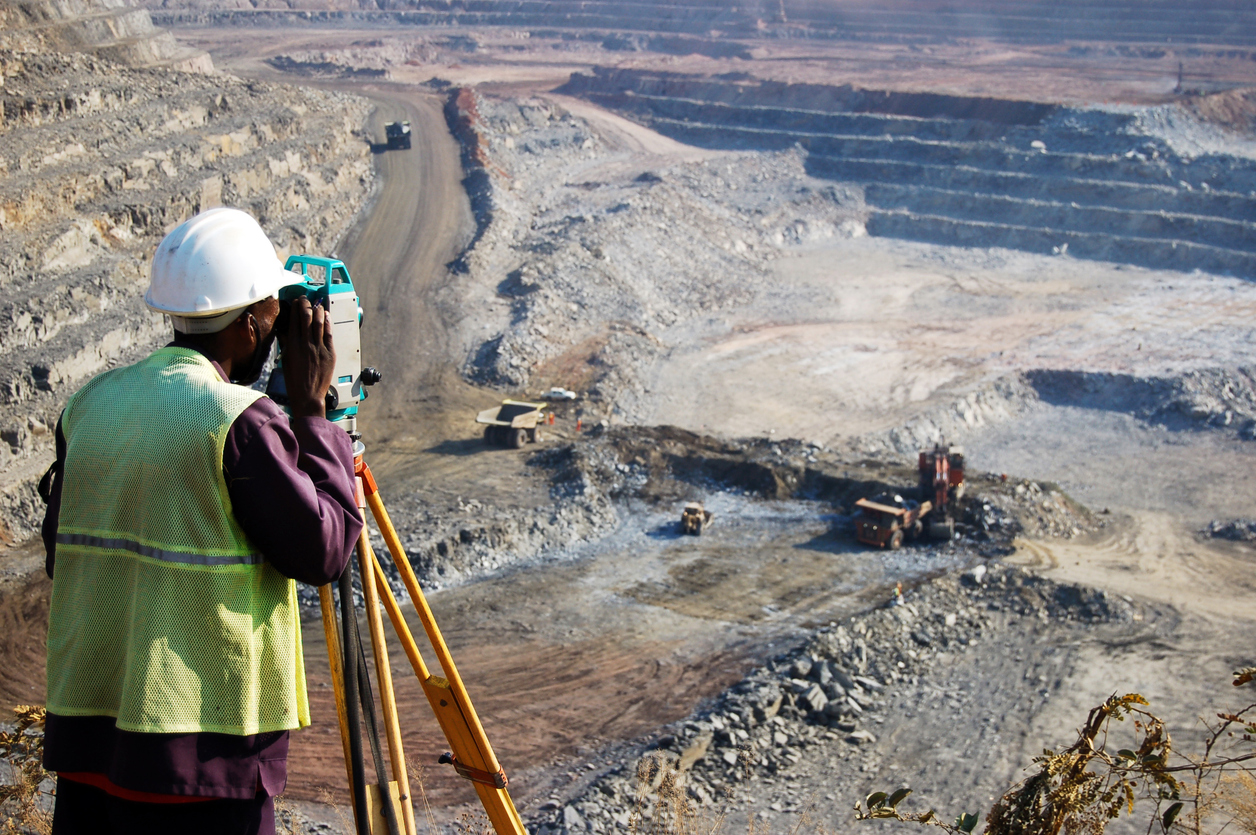 "A locally employed surveyor at an open-pit copper mine in Zambia.  He peers through his survey instrument to record the daily changes in the open-pit, and help guide mining activities to the engineer's plans."
